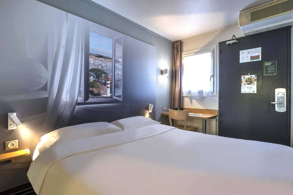 A hotel room with a bed, a desk, a chair, a window with curtains, and a door with a calendar and a sign.