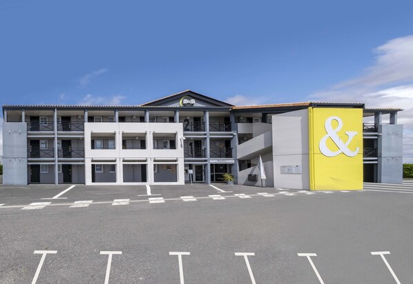 A modern building with a prominent yellow sign featuring a white ampersand symbol. It has multiple floors with balconies and a central entrance.