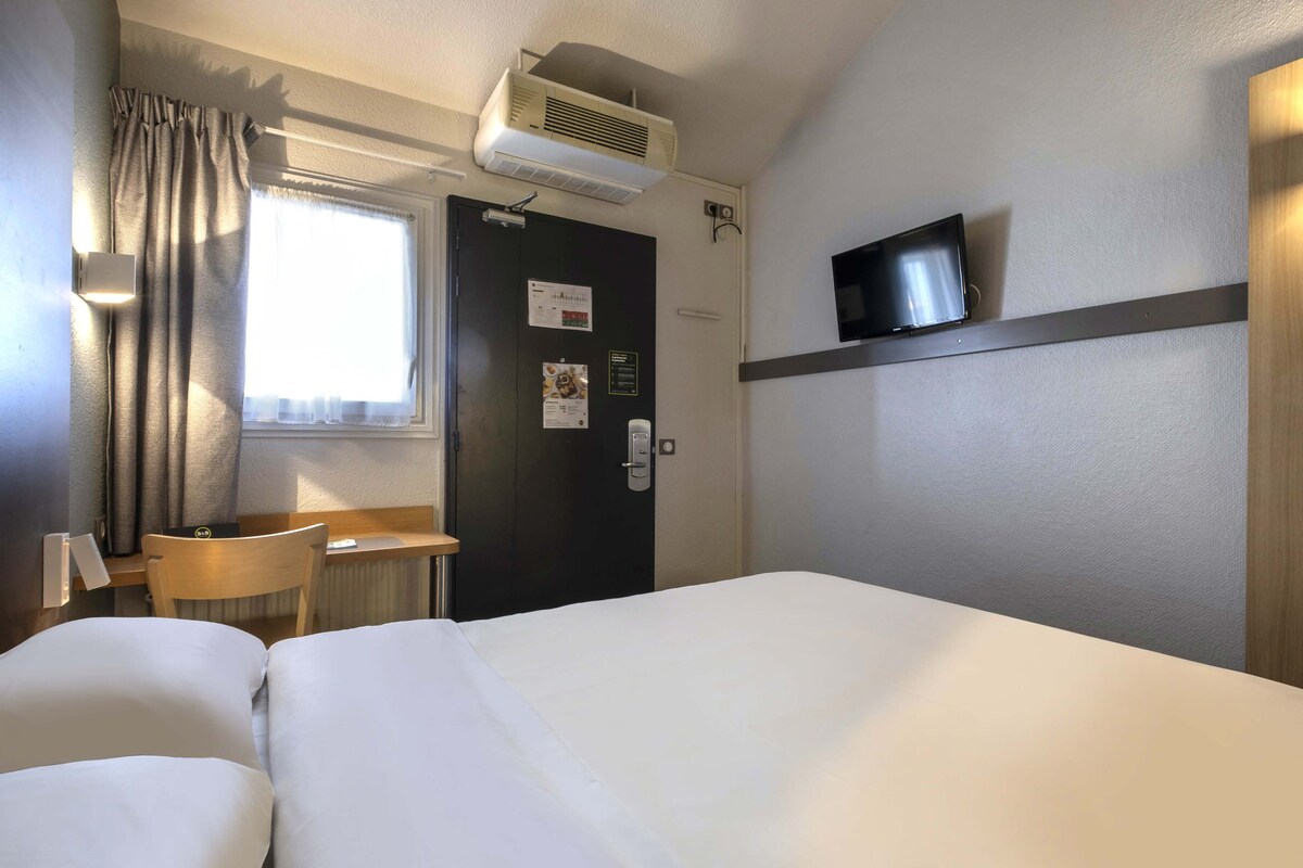 A hotel room with a bed, a desk, a chair, a TV, and an air conditioning unit.