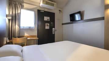A hotel room with a bed, a desk, a chair, a TV, and an air conditioning unit.