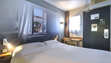 A hotel room with a bed, a desk, a chair, a window with curtains, and a door with a calendar and a sign.