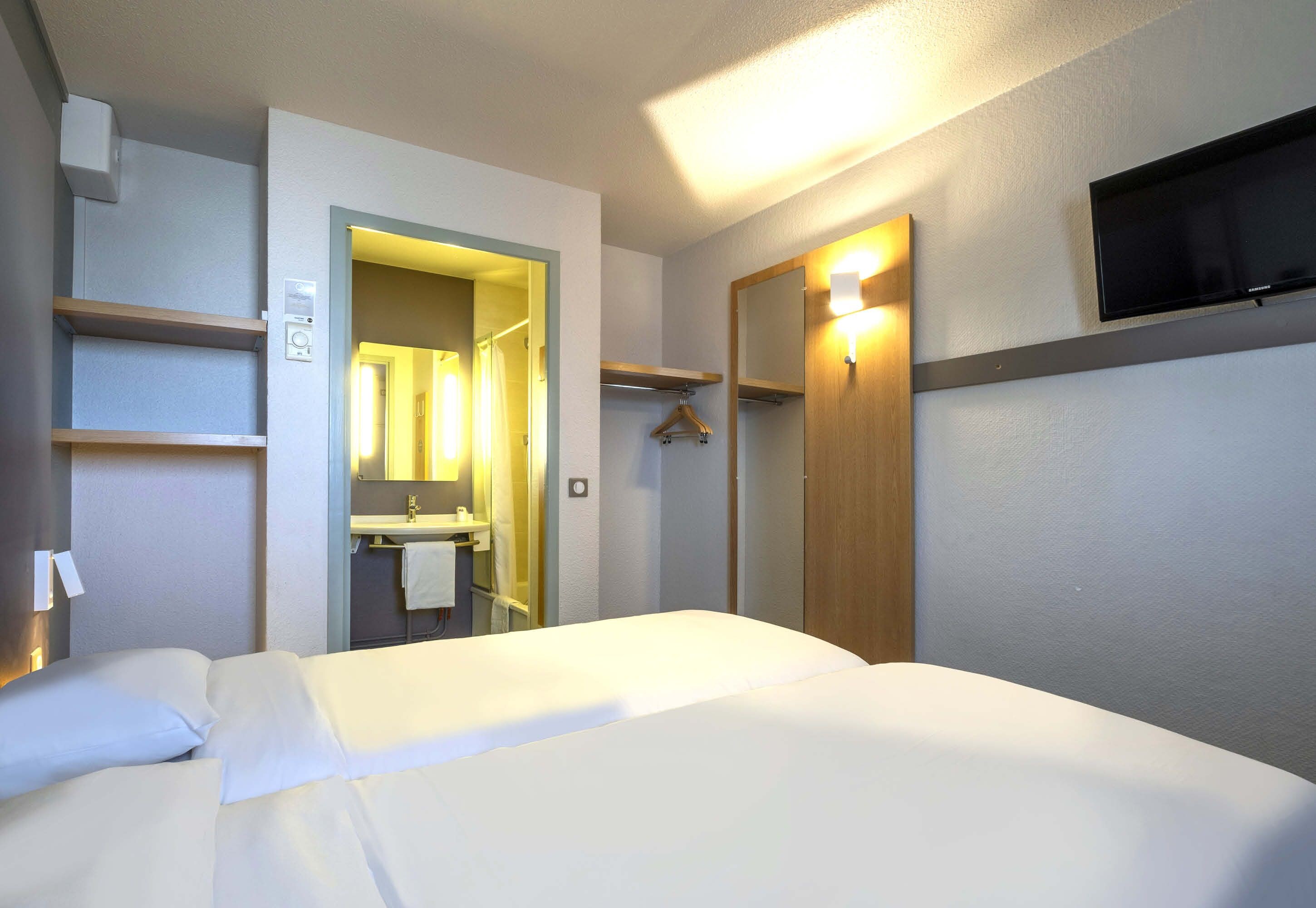 A hotel room with a bed, a mirror, a TV, and a bathroom visible through an open door.