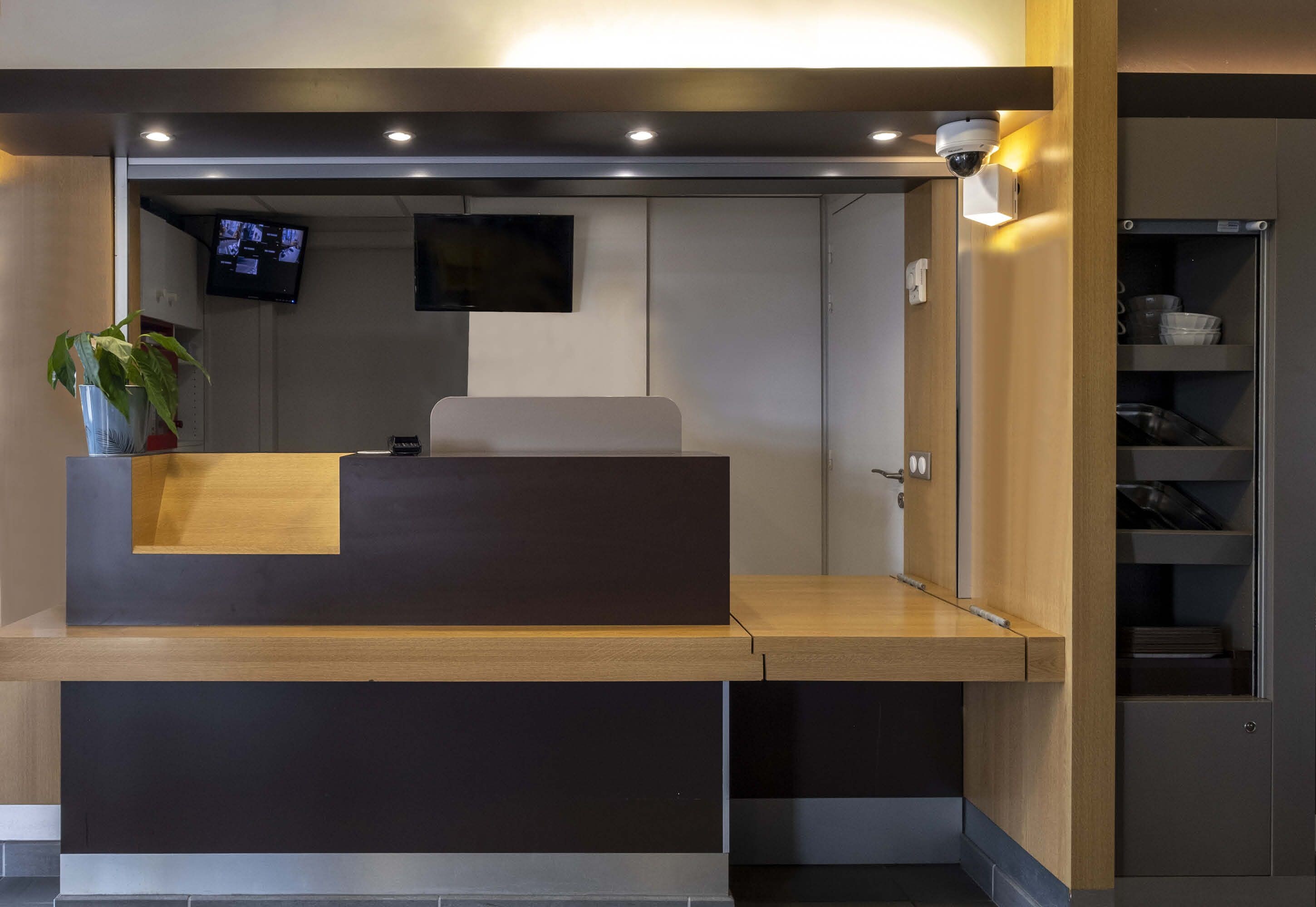 A reception desk with a flat-screen TV mounted above it, a potted plant to the left, and a wooden counter.