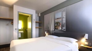Quarto de hotel com uma cama grande, uma janela com vista para edifícios, uma porta que dá para um banheiro e uma mesa de cabeceira com uma luminária.