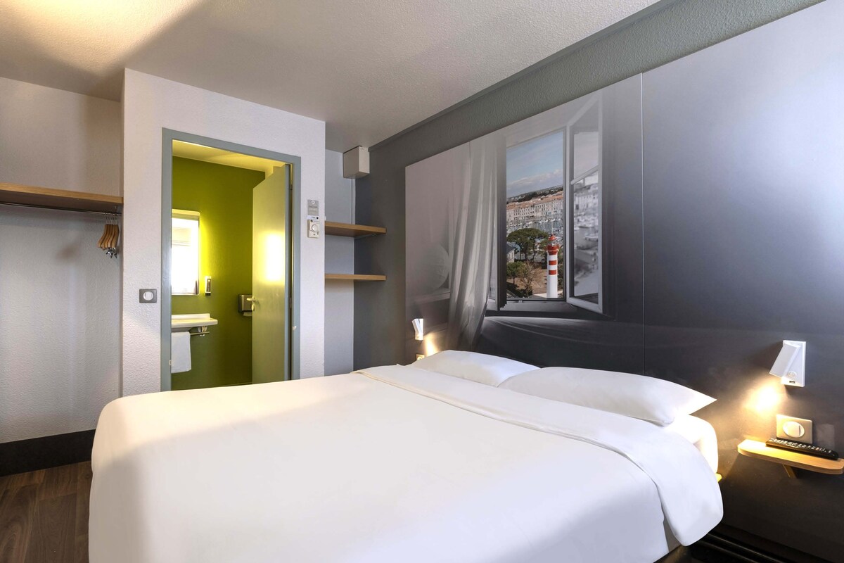 A hotel room with a large bed, a window with a view of buildings, a door leading to a bathroom, and a bedside table with a lamp.