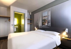 A hotel room with a large bed, a window with a view of buildings, a door leading to a bathroom, and a bedside table with a lamp.