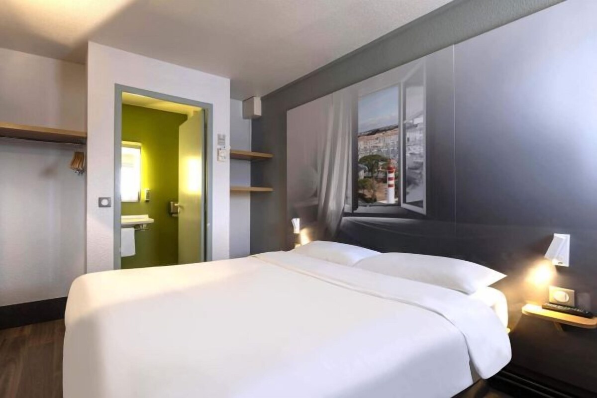 A hotel room with a bed, bedside lamps, a window with curtains, a mirror, and a door leading to a bathroom.