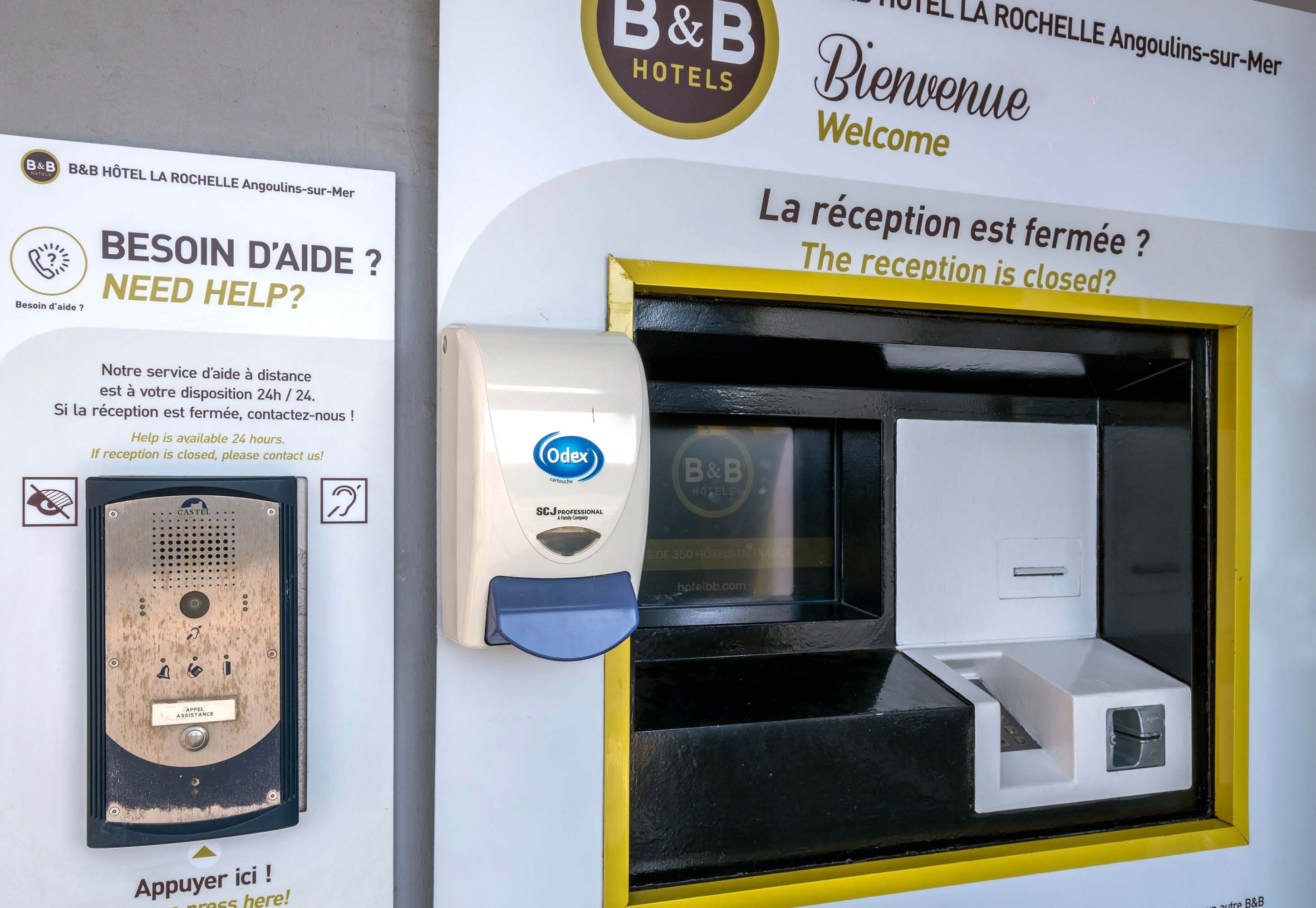 Zona de recepción de hotel con un quiosco de autoayuda, mostrador de recepción y un cartel que indica que la recepción está cerrada.