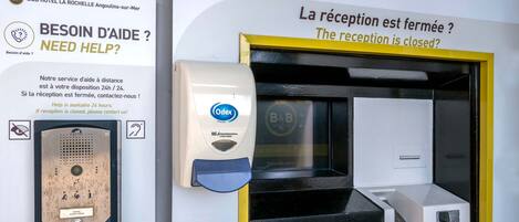 Uma área de recepção de hotel com um quiosque de autoatendimento, balcão de recepção e um aviso indicando que a recepção está fechada.
