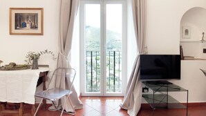 A room with a TV on a glass table, a chair, a table with fruits and a vase, and a painting on the wall. - Casa Vacanza Eos (Sperlonga)
