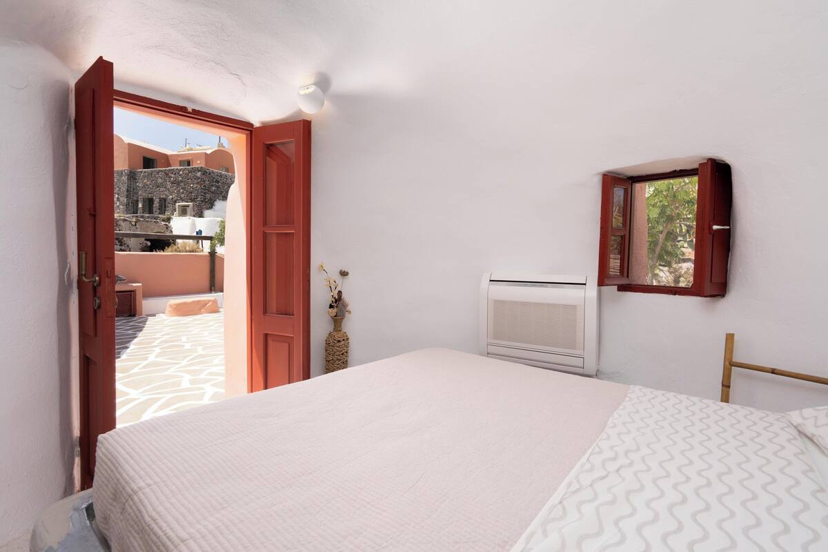 Een slaapkamer met een bed, een airconditioner, een klein tafeltje met een vaas en uitzicht op een binnenplaats door een geopende deur.