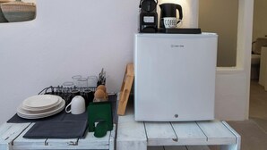 Cocina compacta con un refrigerador blanco, una cafetera y una taza en la encimera. Hay una pequeña mesa de madera con platos y un juego de cuchillos.