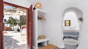 Una habitación con paredes blancas, una puerta de madera, una estantería con ropa, una mesita y vistas a edificios.