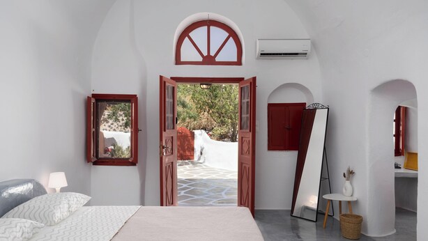 Una habitación con cama, un espejo, una mesita con un florero y una puerta abierta que da a un espacio exterior con suelo de piedra y vegetación.