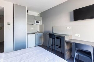 Kompakti hotellihuone, jossa on sänky, pieni keittiö, työpöytä ja seinälle asennettu televisio.