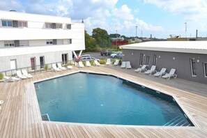A swimming pool with a wooden deck, surrounded by lounge chairs and a building with multiple windows. - Residence le Terral - Vacancéole (Saint-Jean-de-Vedas)