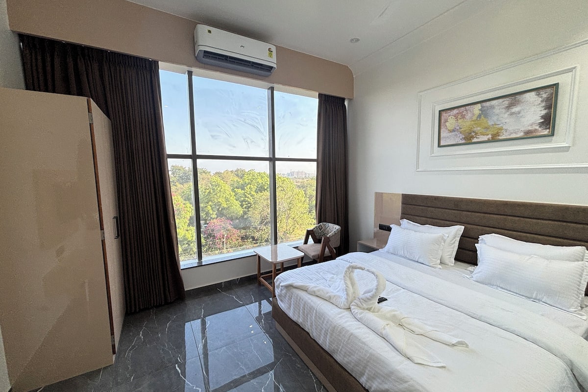 A hotel room with a large bed, a desk, and a chair. There is a window with curtains, an air conditioning unit, and a painting on the wall.