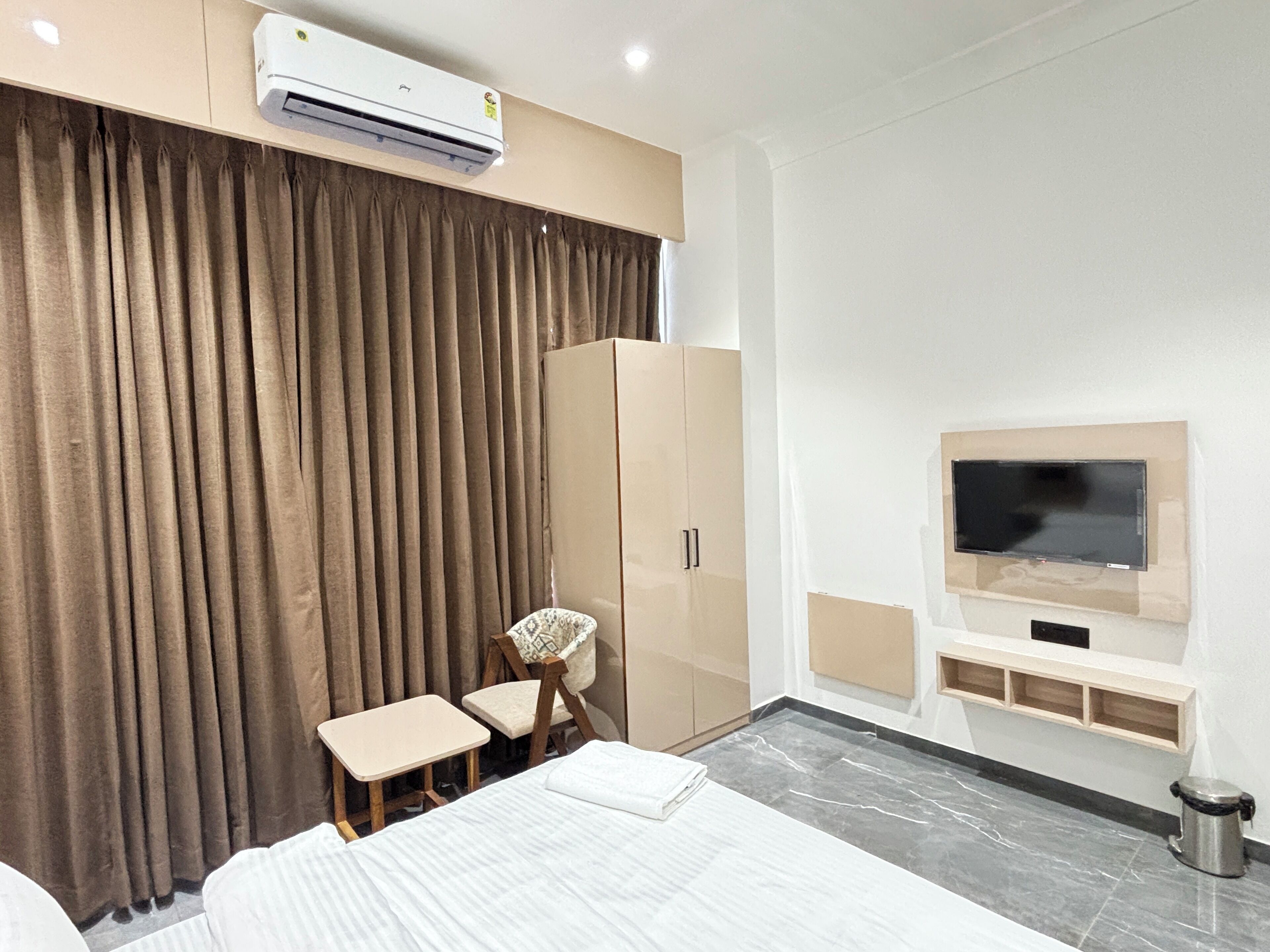 A modern hotel room with a large bed, a small table, a chair, a wardrobe, an air conditioning unit, a television, and a trash bin.