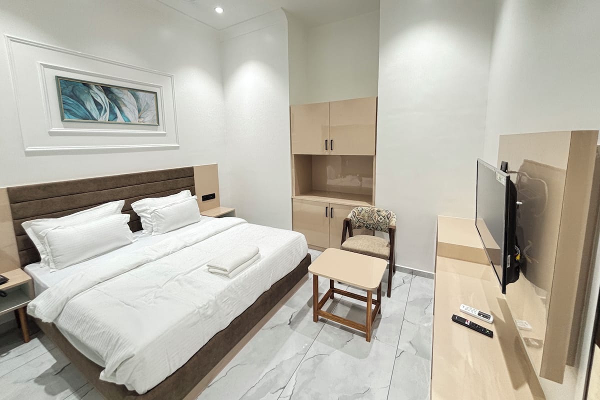 A modern hotel room with a large bed, bedside tables, a small wooden stool, a TV, and built-in cabinets.