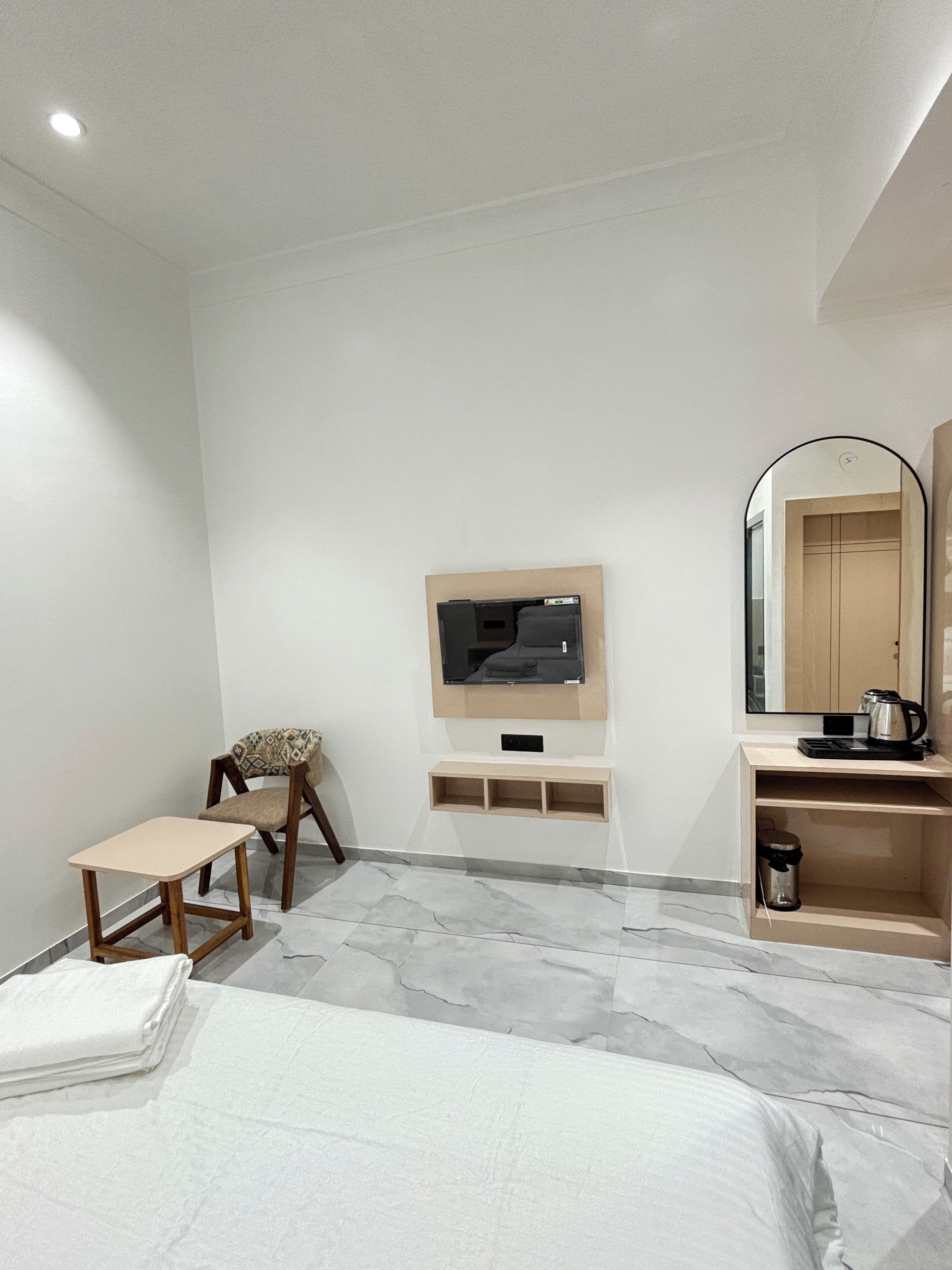 A hotel room with a bed, a TV mounted on the wall, a wooden chair, a small table, a shelf with a kettle, and a mirror.