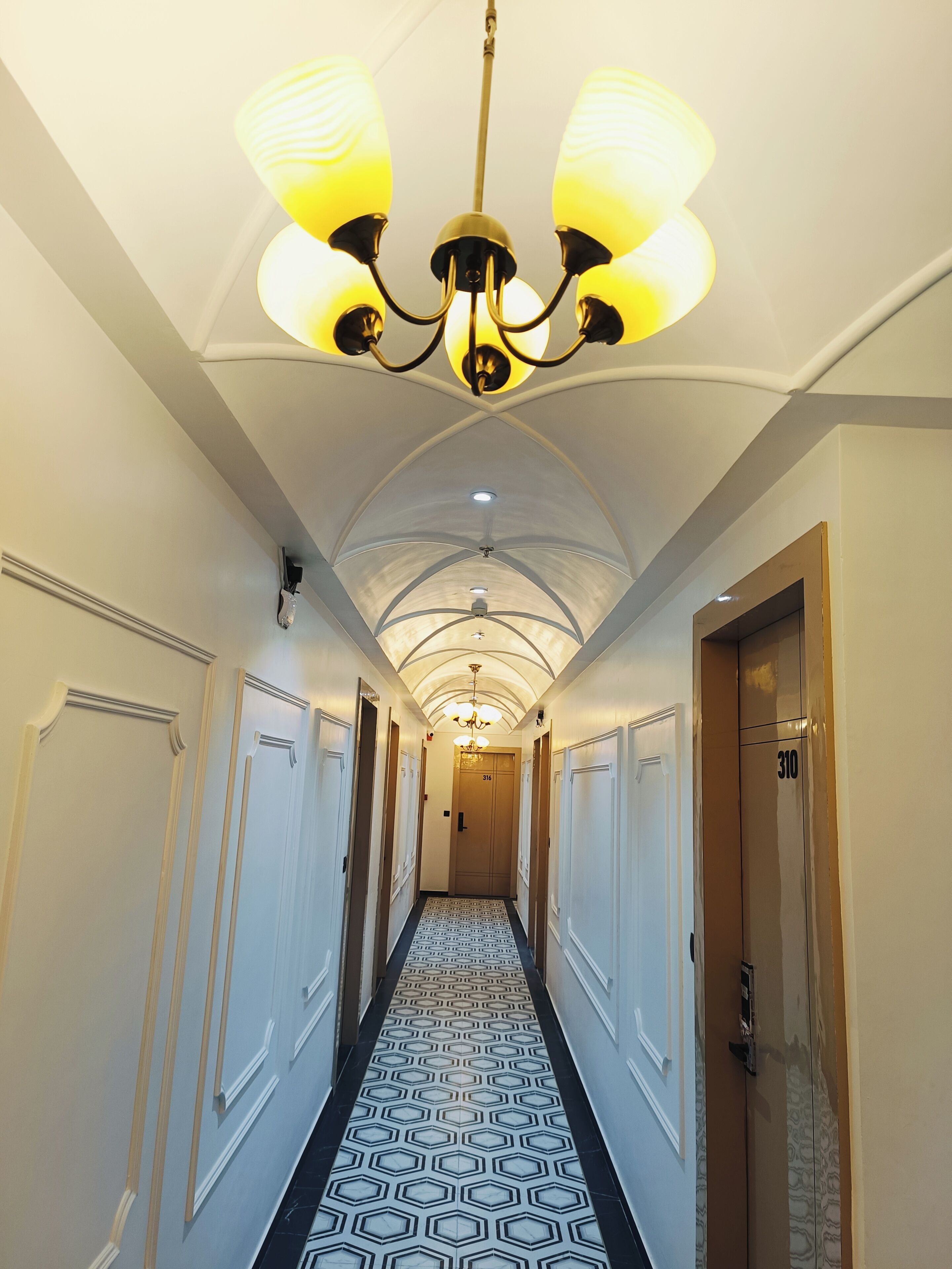 Un pasillo de hotel con piso estampado, un candelabro y puertas con los números 316 y 310.