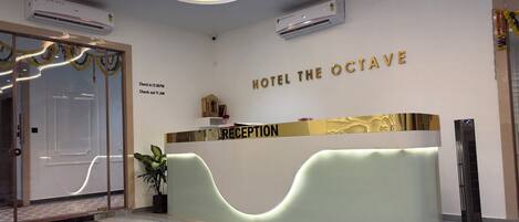 A hotel reception area with a modern design, including a reception desk, air conditioning units, and a check-in/out schedule on the wall.