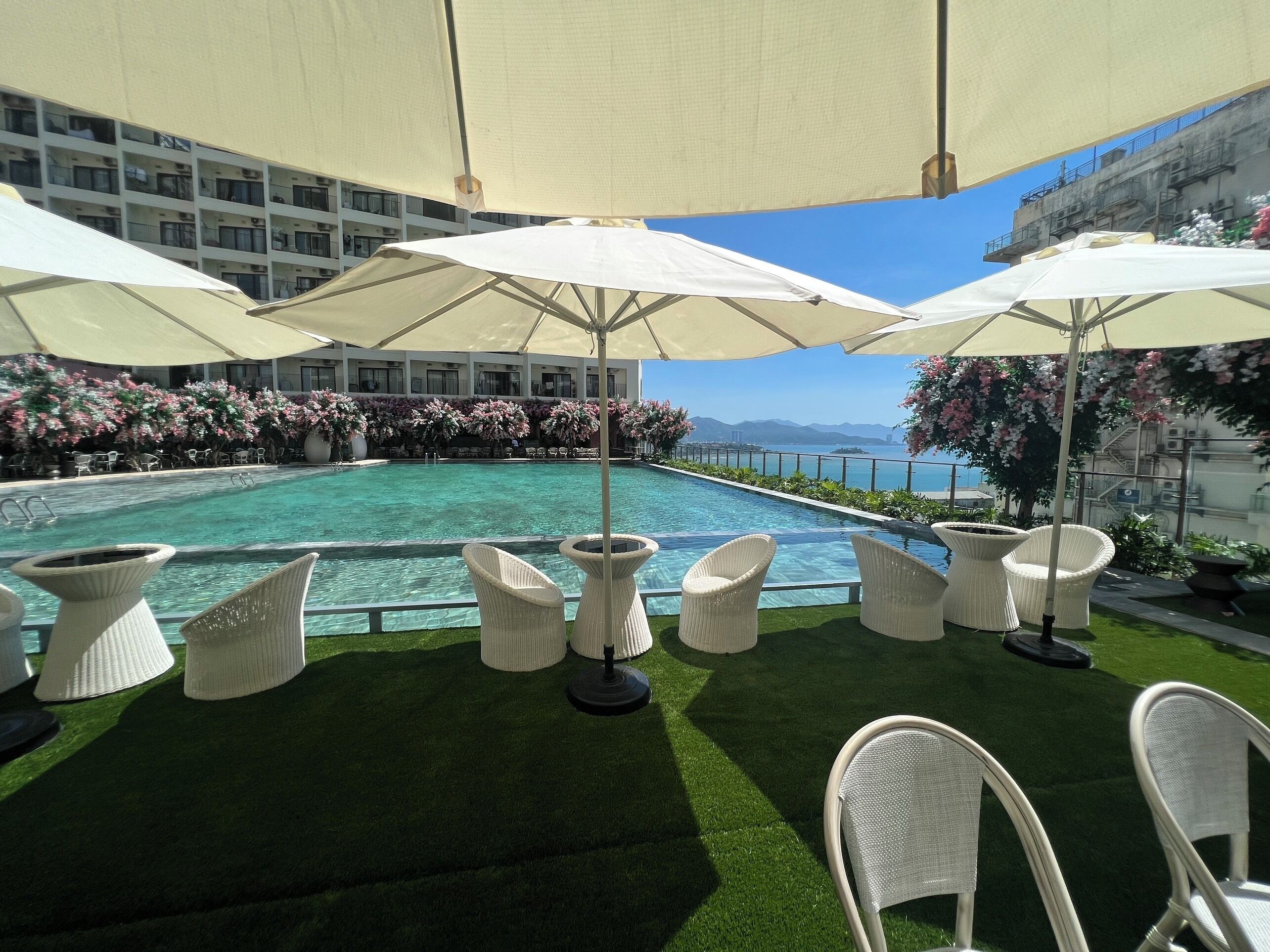 A poolside area with white wicker chairs and umbrellas, surrounded by a pool and a view of the sea.