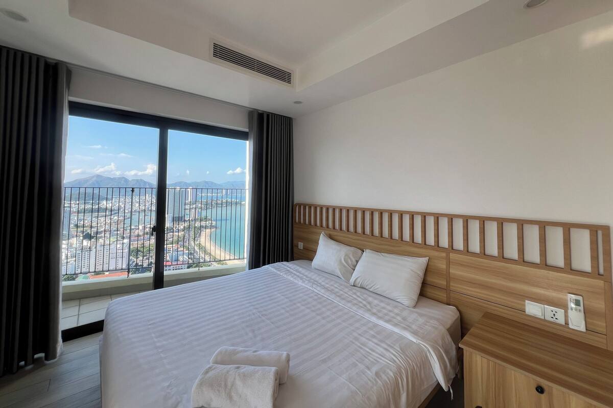 Habitación de hotel moderna con una cama grande, cabecera de madera y vistas a la ciudad a través de ventanas de techo a piso.