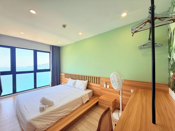 Habitación de hotel con cama, escritorio, una silla, un ventilador de pie y vista al mar a través de ventanas amplias.
