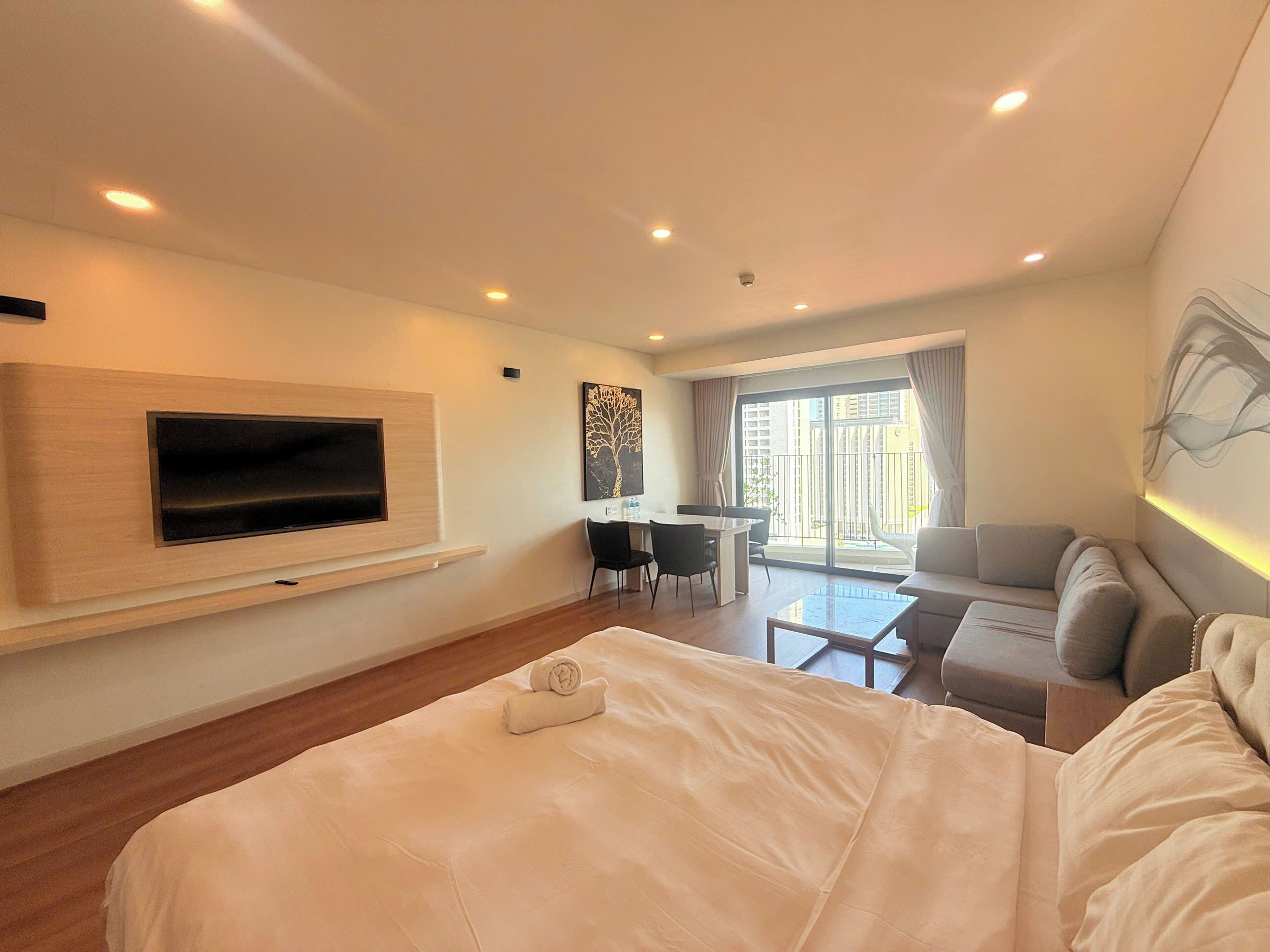 Una habitación de hotel moderna con una cama grande, un televisor de pantalla plana montado en la pared, un rincón de comedor con mesa y sillas, y un balcón con vistas.