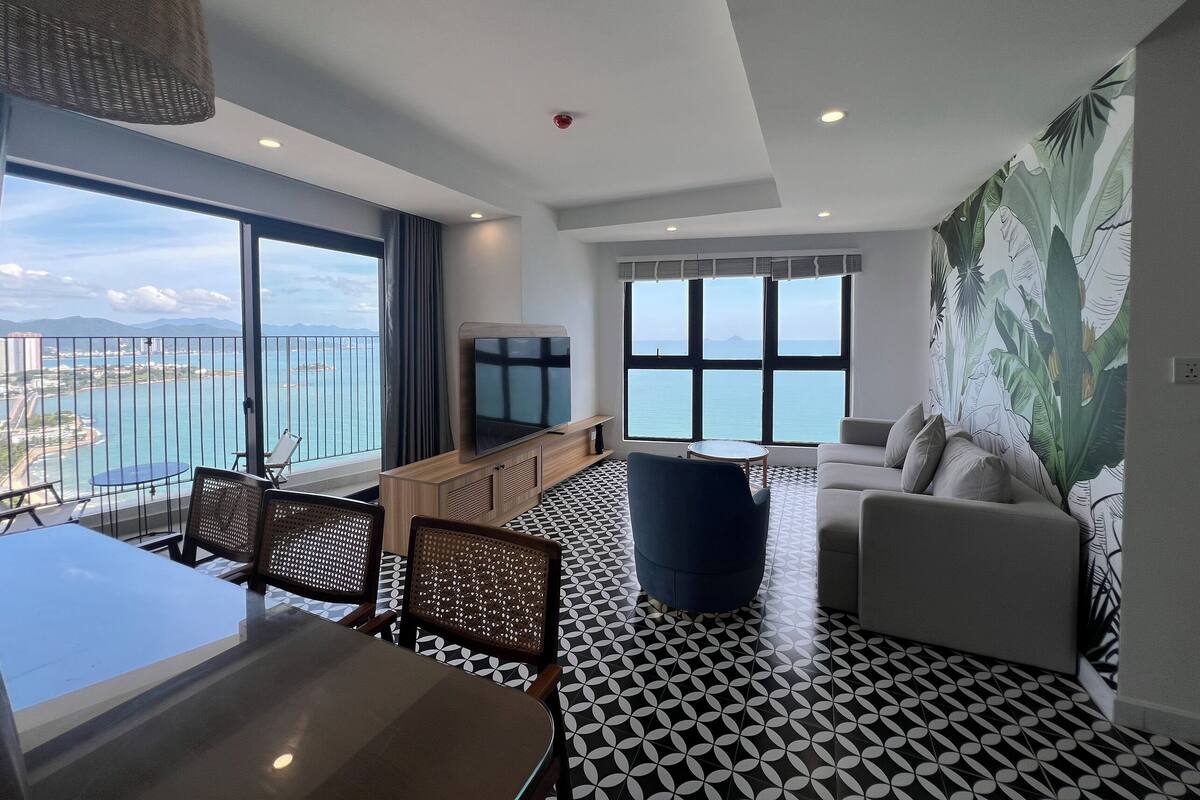 Un salón moderno con un amplio ventanal que ofrece vistas al mar, una zona de comedor con suelo estampado, y un área de estar con un sofá y sillones.