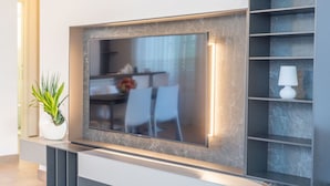 Un salon moderne avec un grand téléviseur à écran plat fixé sur un mur en marbre, une plante en pot et une petite table avec des chaises.