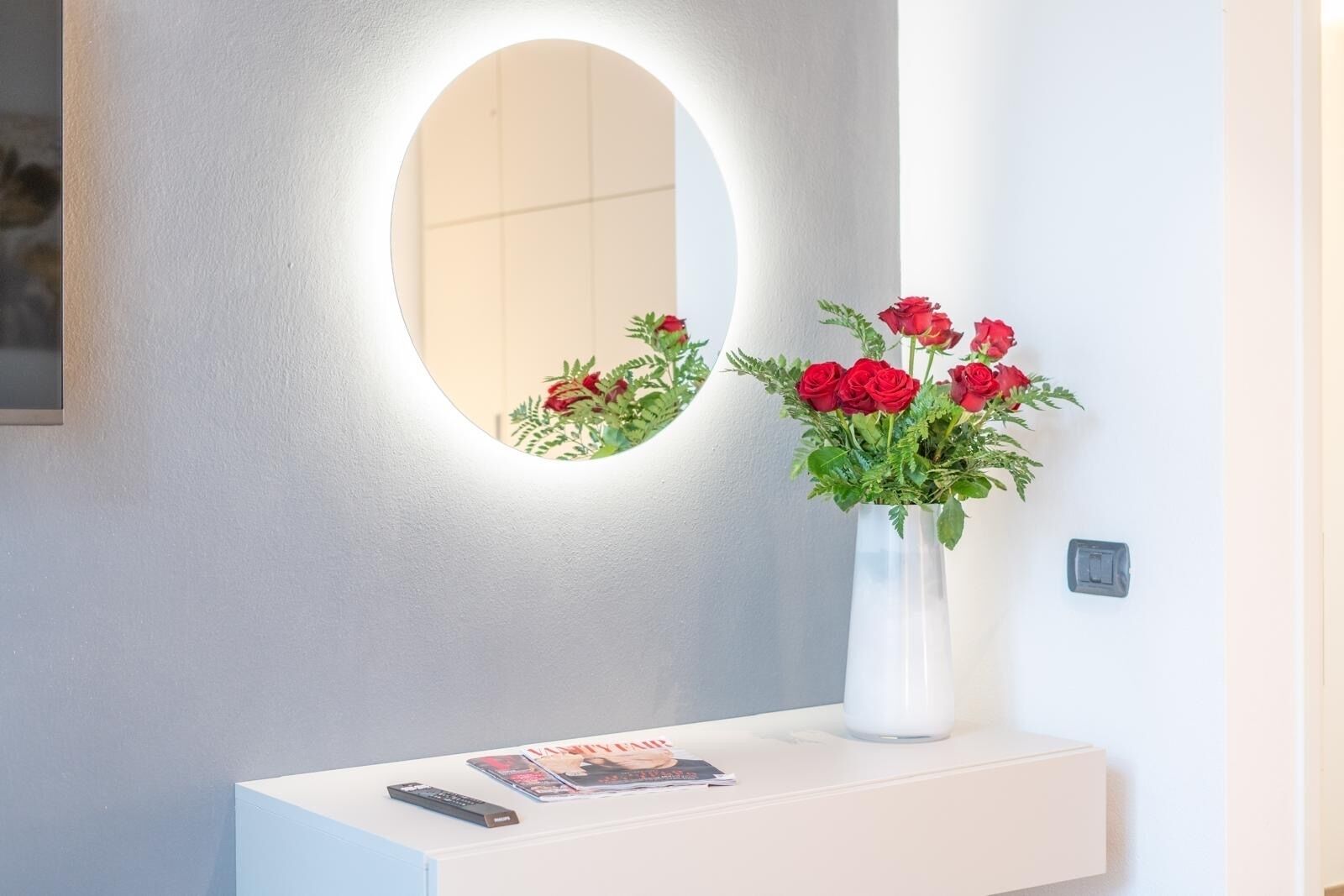 Een witte console tafel met een afstandsbediening en een tijdschrift, een witte vaas met rode rozen, en een spiegel met verlichting, bevestigd aan de muur.
