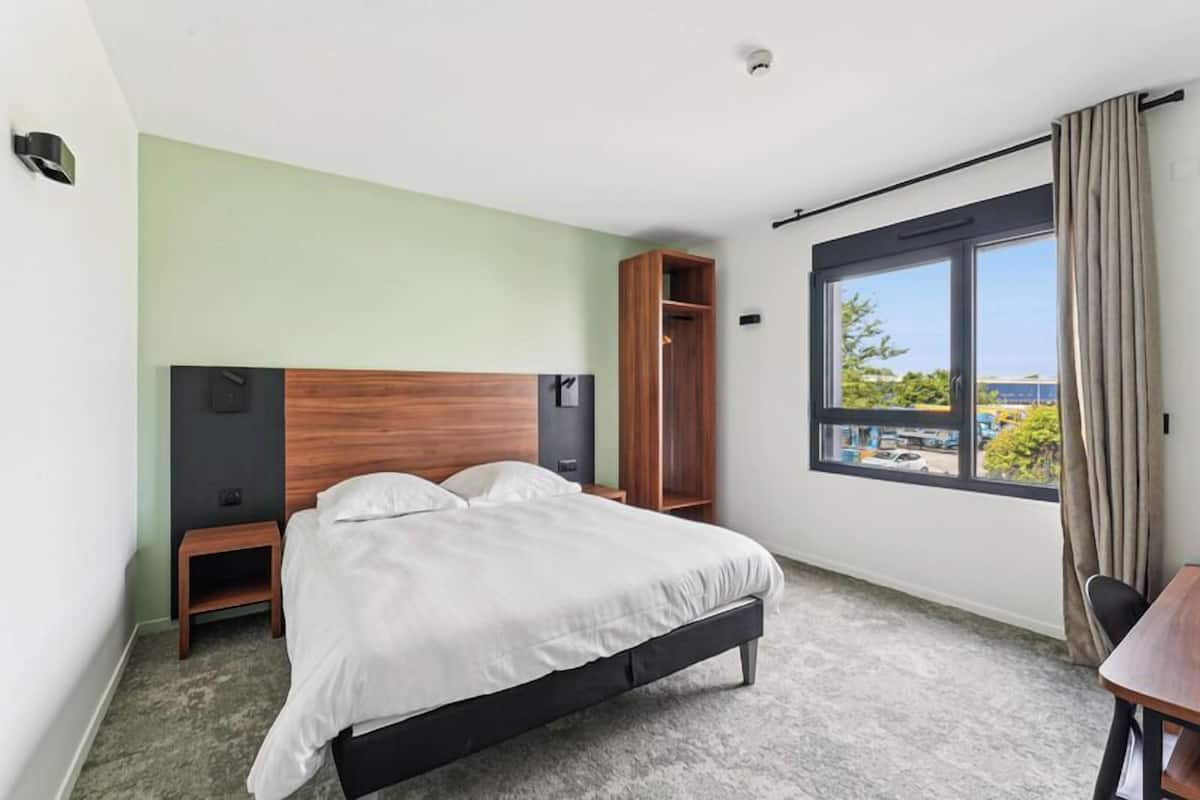 A hotel room with a bed, a wooden wardrobe, a small table, and a window with a view of the outside.