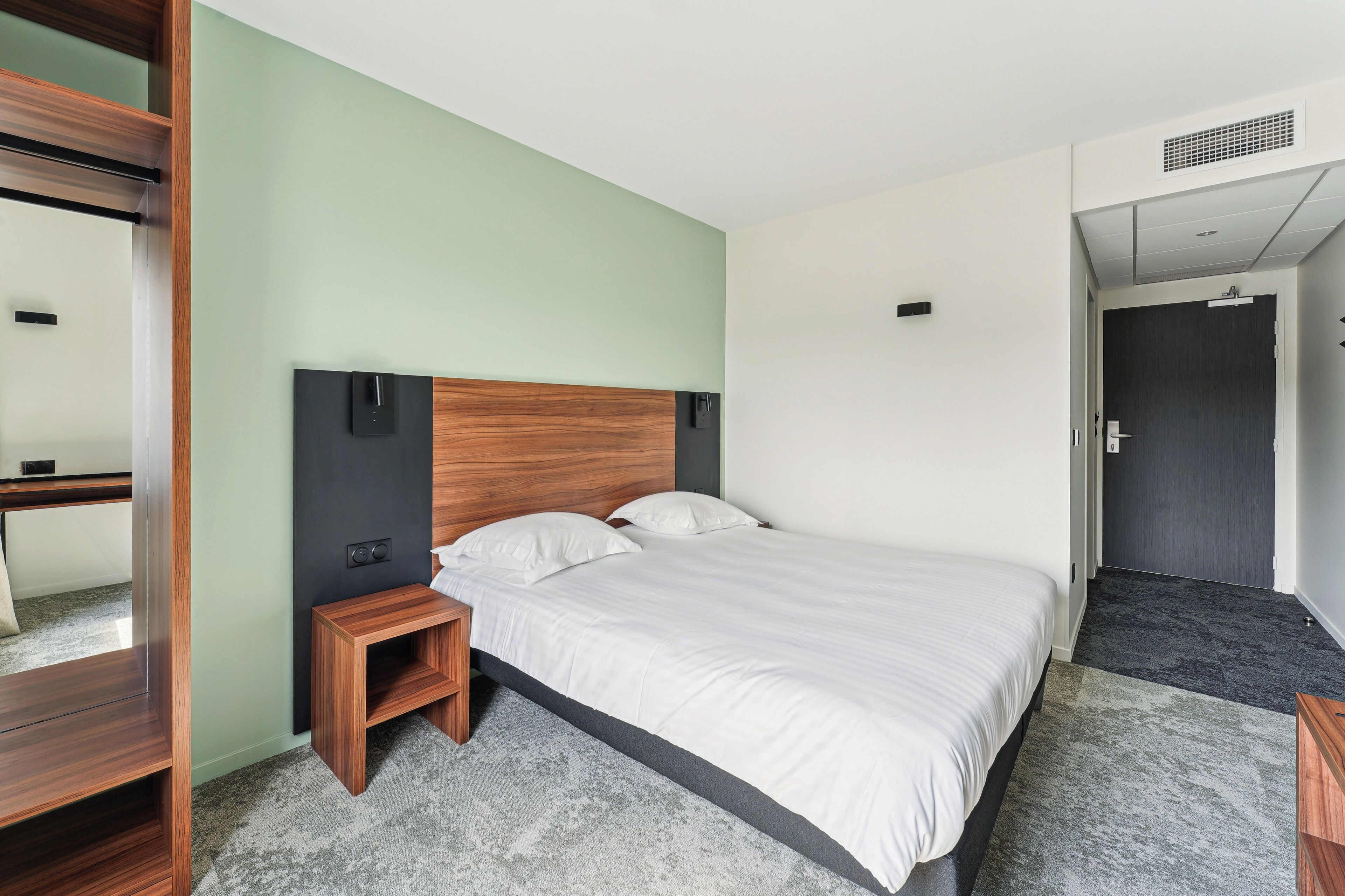 A modern hotel room with a bed, a nightstand, a wooden wardrobe, and a door leading to another room.