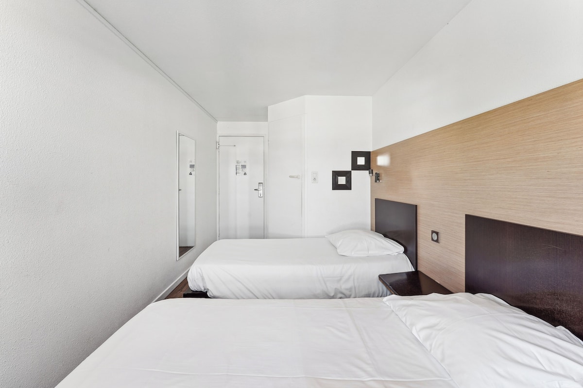 A modern hotel room with two single beds, a wooden headboard, a large mirror, and a door leading to a bathroom.