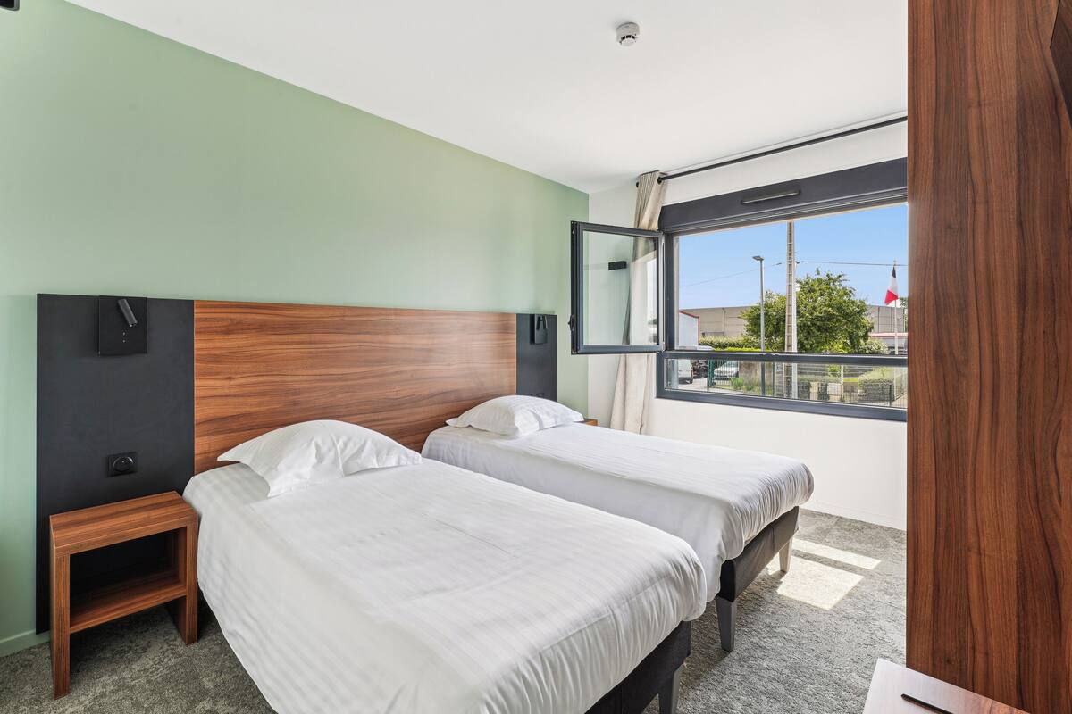 A hotel room with two single beds, a wooden headboard, a nightstand, and a large window with a view of the outside.