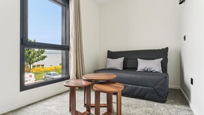 Een kamer met een grijze bank, twee houten krukken, een groot raam met uitzicht op een geel gebouw en een auto, en een wit tapijt.