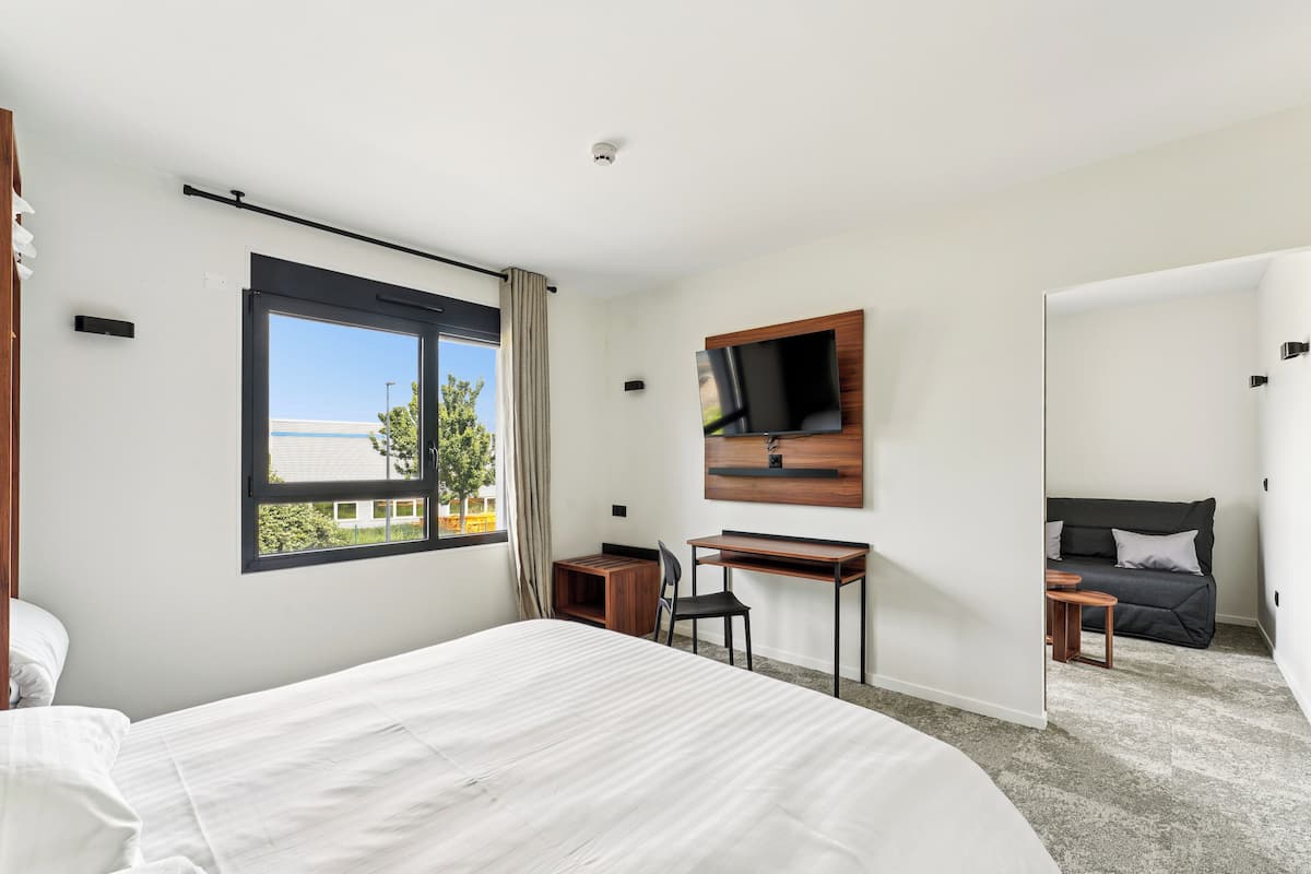 A modern hotel room with a large bed, a desk, a TV mounted on the wall, and a view of the outside.
