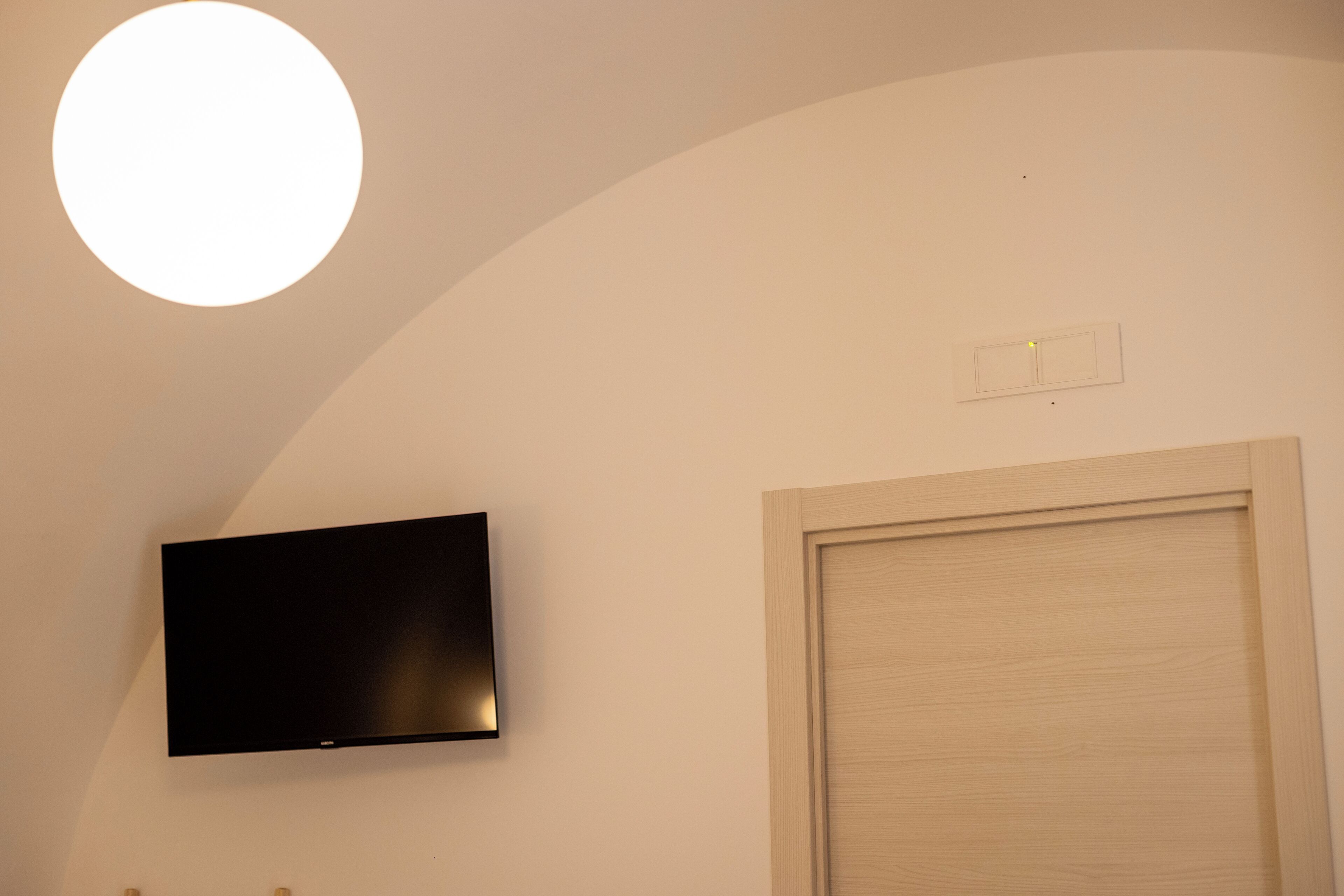 A room with a flat-screen TV mounted on the wall, a wooden door, and a ceiling light fixture.