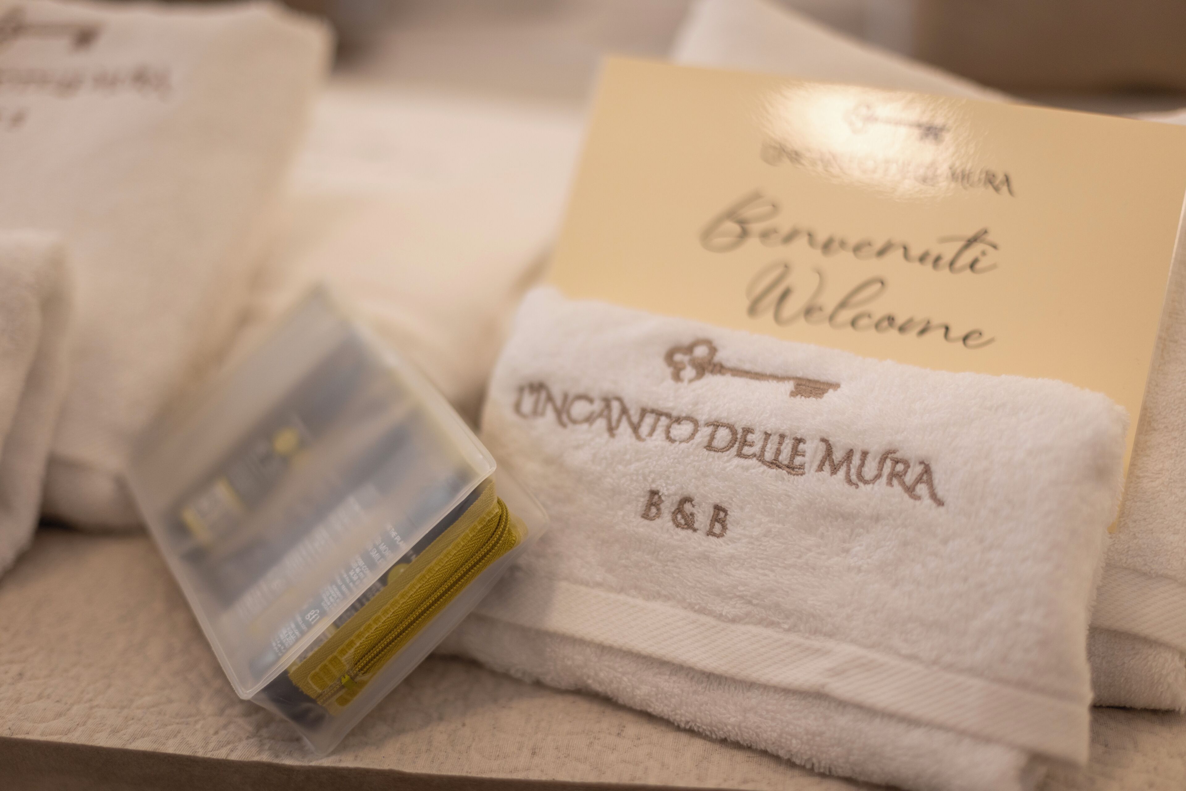 A towel with 'Benvenuti Welcome' and 'Uncanto delle Mura B&B' embroidered on it, alongside a small plastic container.