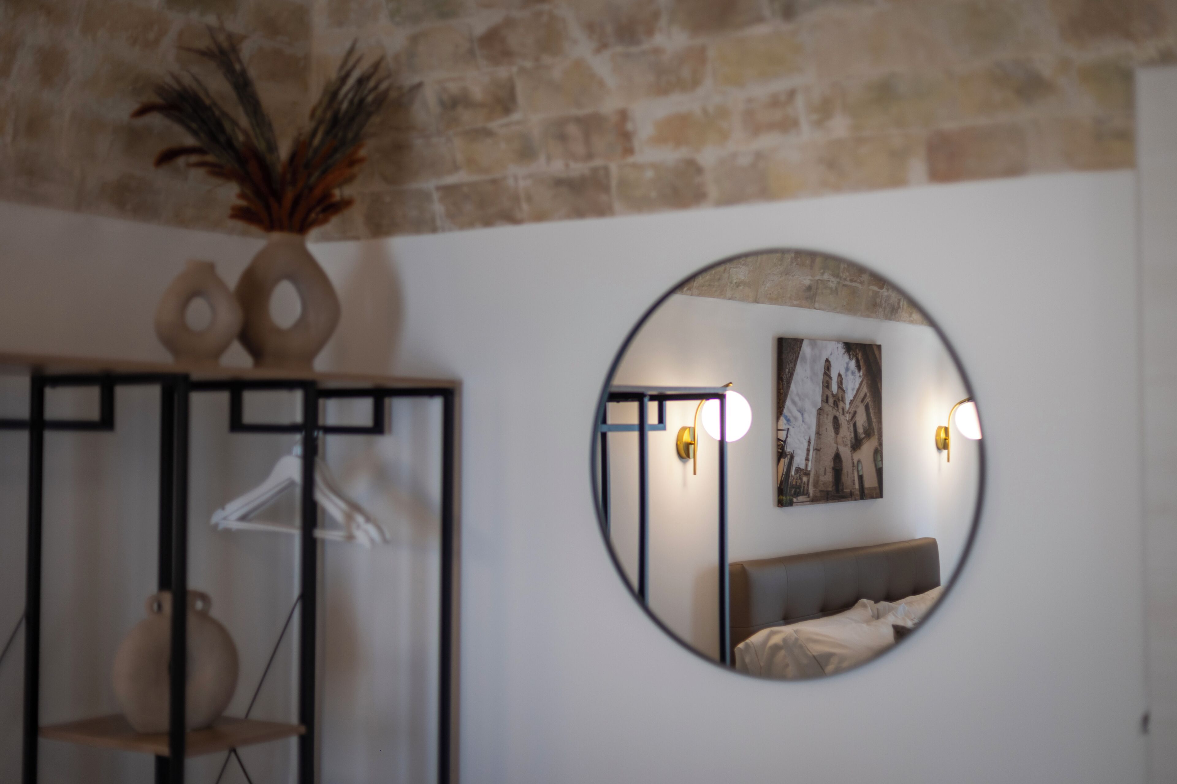 A round mirror reflecting a bedroom with a bed, wall-mounted lights, and a painting.