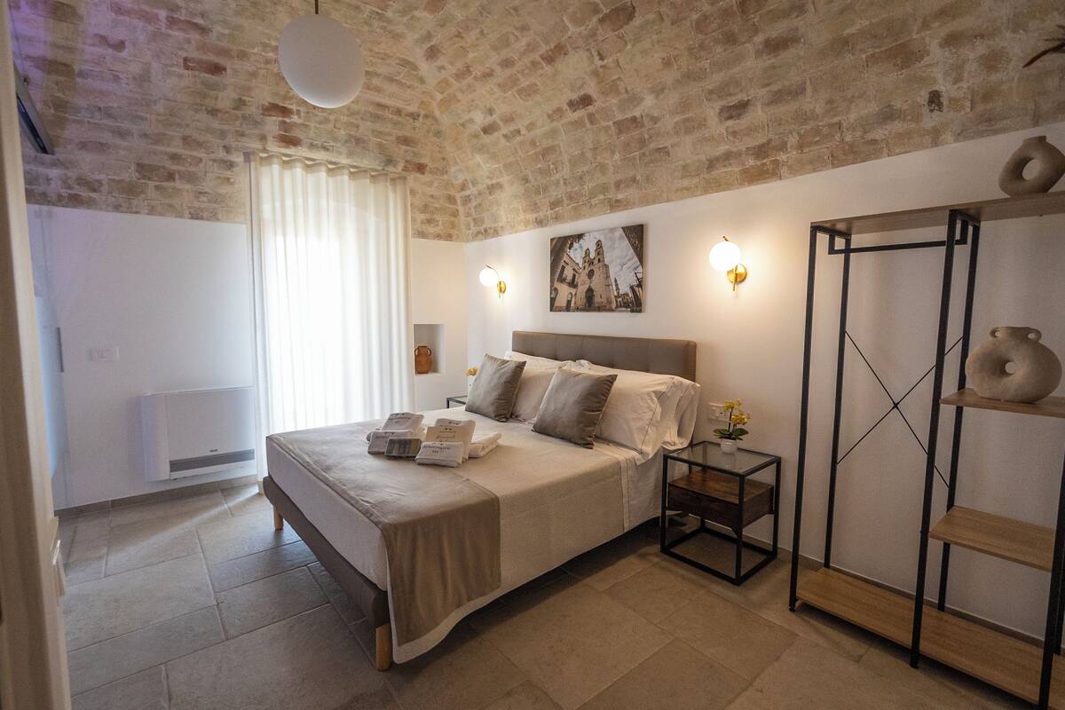 A bedroom with a brick ceiling, a bed with a beige bedspread, a bedside table with a lamp, and a wall-mounted shelf with decorative items.