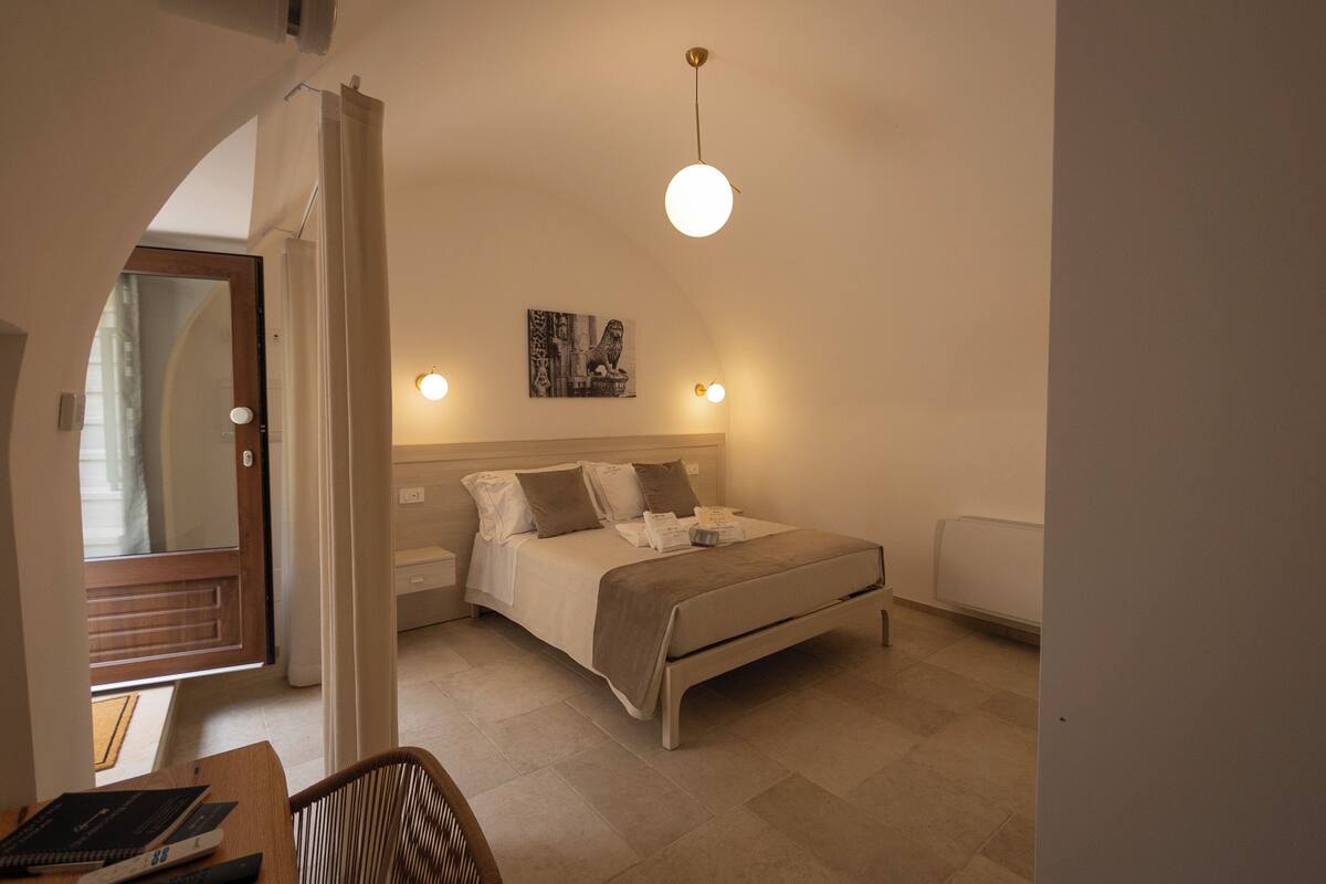 A bedroom with a bed, bedside tables, a hanging light fixture, a painting on the wall, and a door leading to another room.