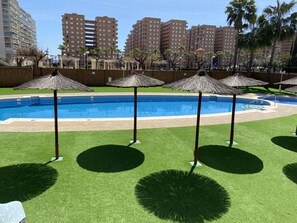 A swimming pool with surrounding grass and palm trees, with tall buildings in the background. - 2º Line Apartment (Oropesa del Mar)
