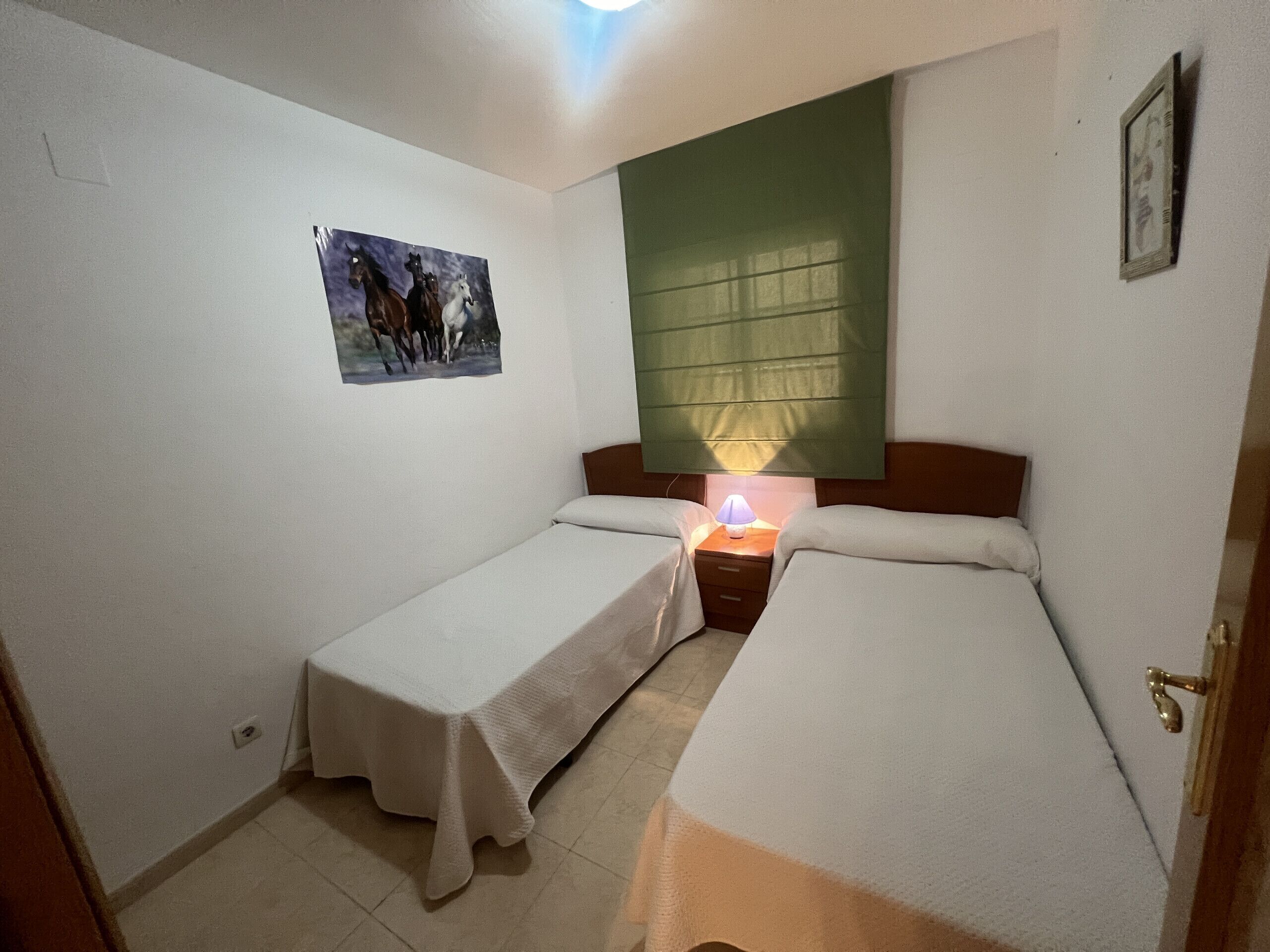 Ein kleines, sauberes Zimmer mit zwei Einzelbetten, einem Nachttisch mit Lampe, einem grünen Fensterrollo und einem an der Wand hängenden Bild von Pferden.