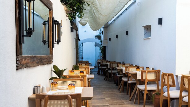 Un espace de restauration en plein air avec des tables et des chaises en bois, des plantes en pot et un mur blanc agrémenté d’un miroir et de lanternes.