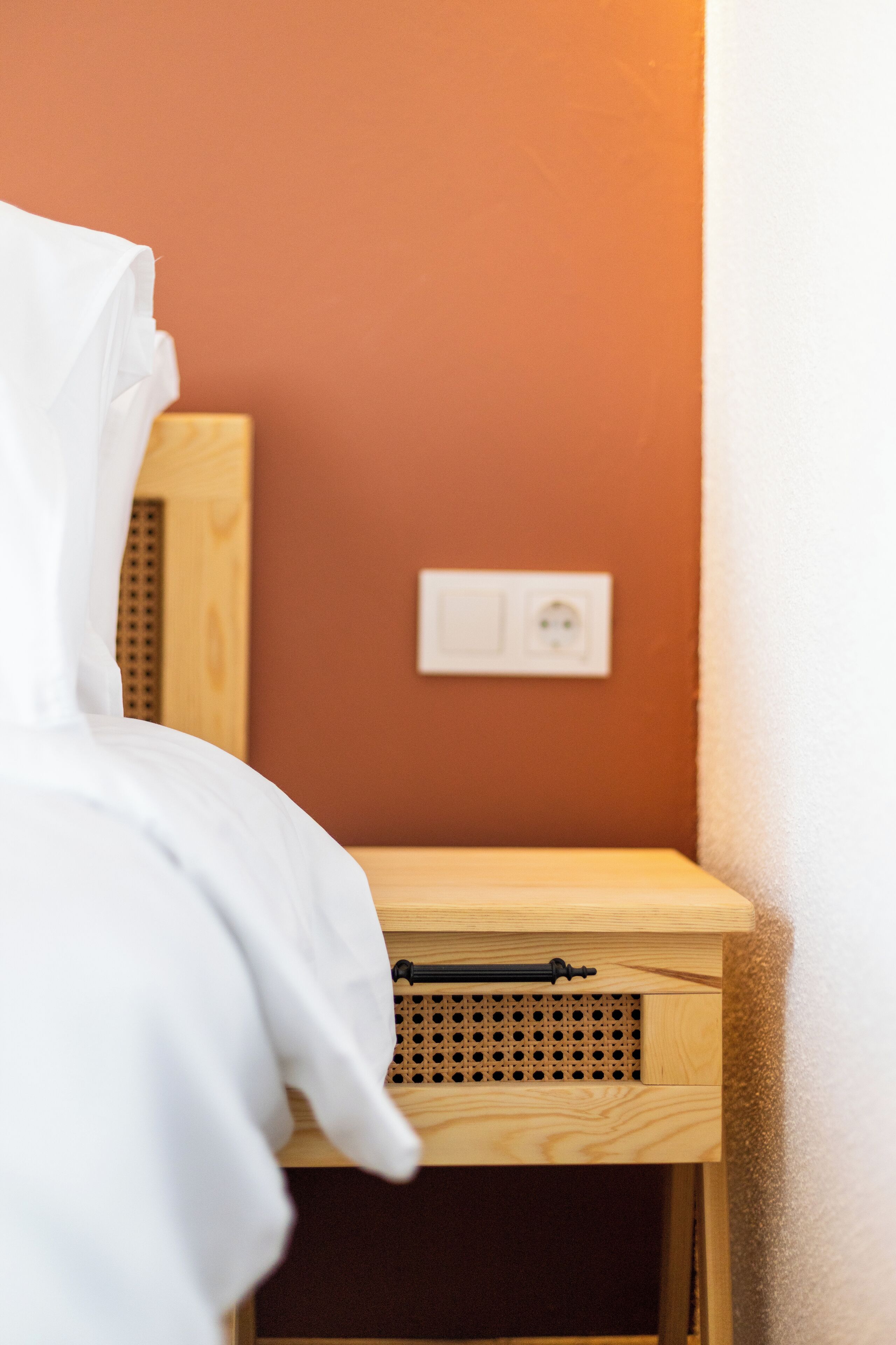 A corner of a room with a wooden nightstand, a white bed, and a wall socket.