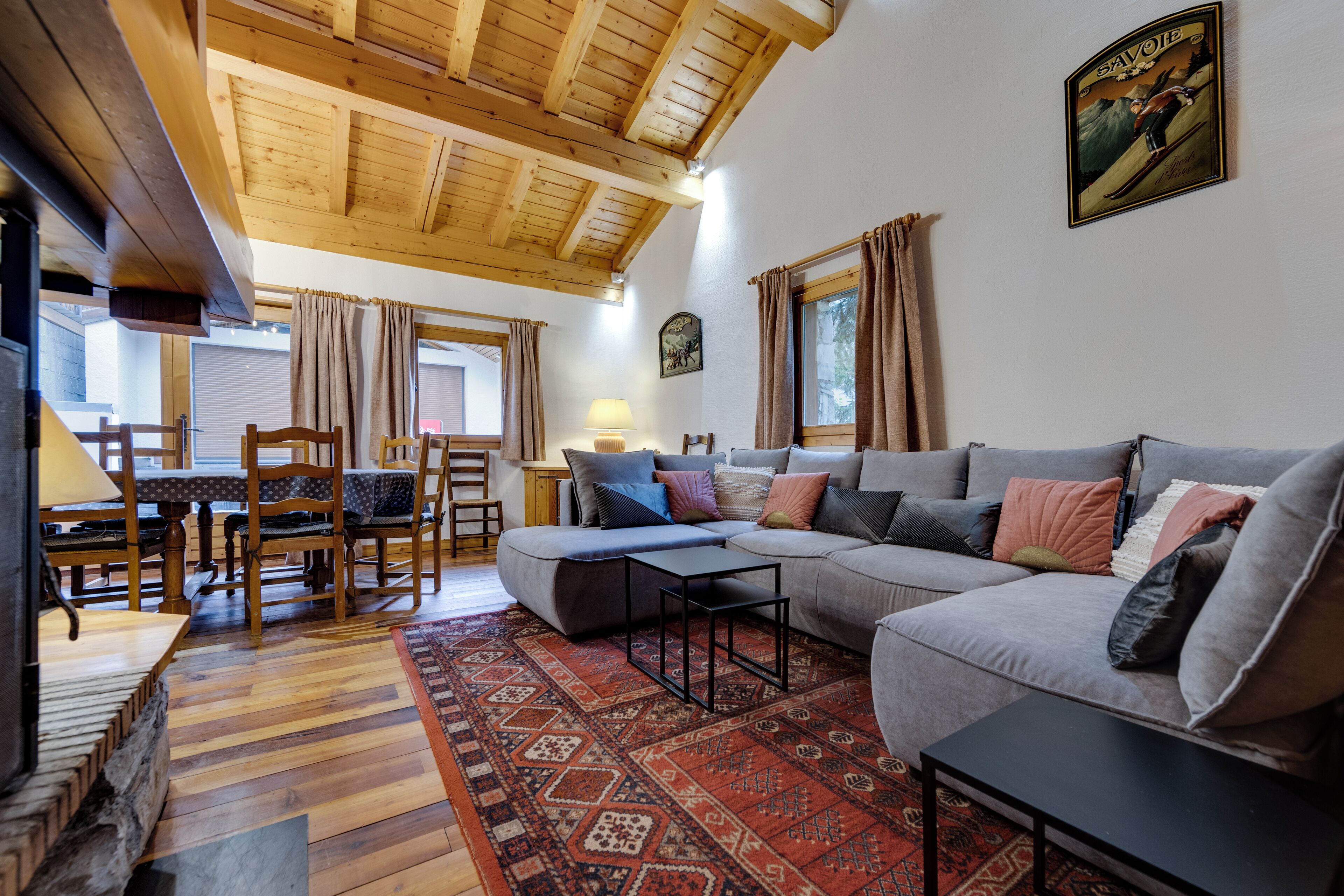 A cozy living room with a wooden ceiling, a grey sectional sofa, a patterned rug, and a dining area with wooden furniture.