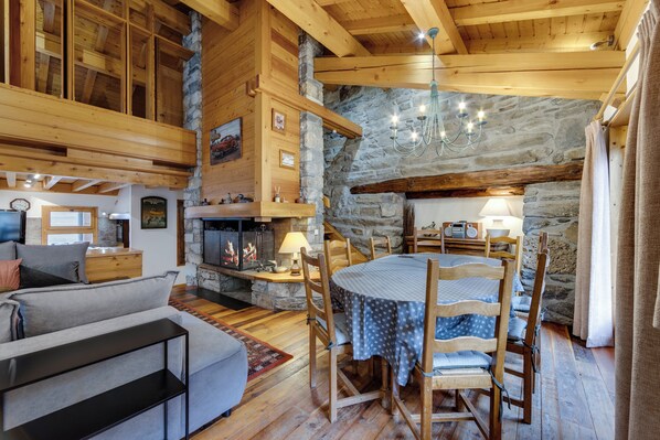 A cozy room with a stone wall, wooden beams, a fireplace, a dining table with chairs, and a sofa.